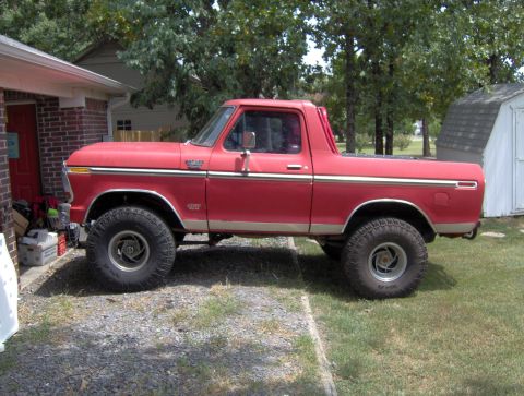 1978 Ford Bronco - A lifted, red 1978 Ford Bronco is parked on a gravel driveway next to a brick building. The vehicle has large off-road tires and a chrome t…