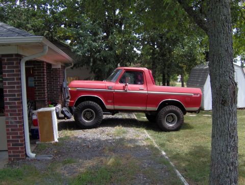 1978 Ford Bronco - A lifted red 1978 Ford Bronco is parked on a gravel driveway next to a brick building and a white shed. The vehicle has large off-road tire…