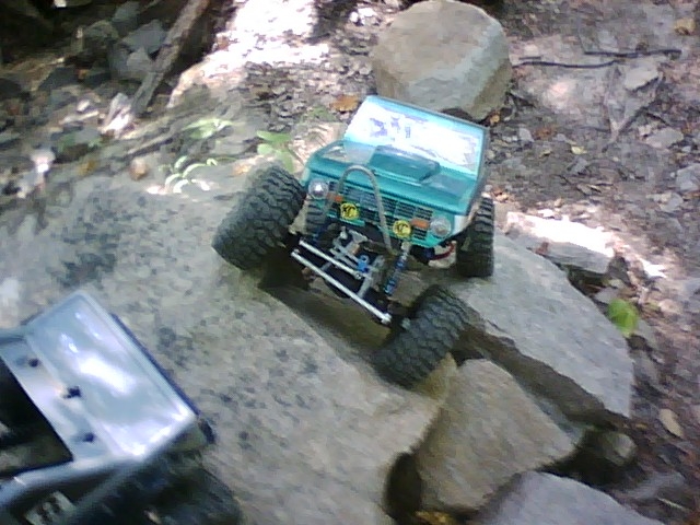 Teal remote-controlled Ford Bronco on rocks with another vehicle part in foreground.