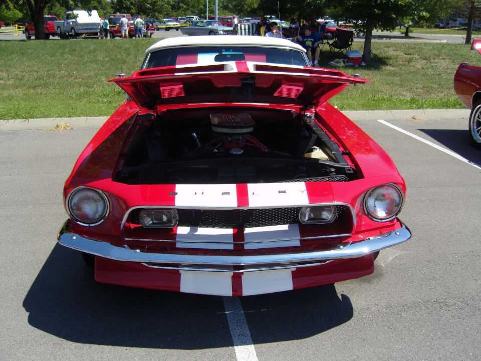 Red classic car with white racing stripes, hood open showing engine. Front view