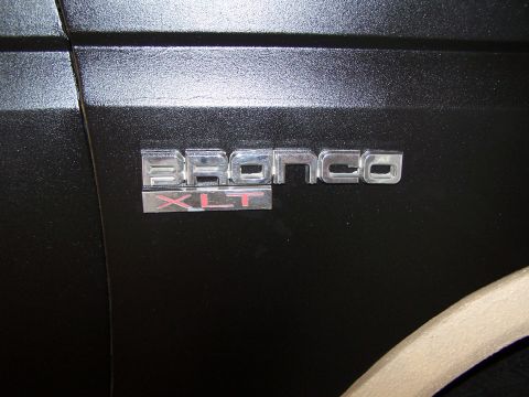 1986 Ford Bronco - Close-up of a Bronco XLT emblem on a dark, textured surface with a portion of a tan wheel visible.