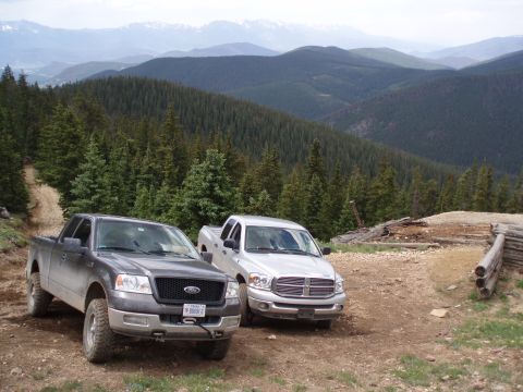 2004 Ford F-150 - Two pickup trucks are parked on a mountain road with a forest and distant mountains behind them.