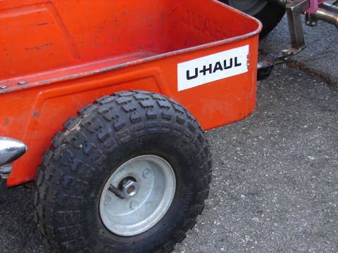1973 Ford Ranchero - Close-up of the rear corner of a small orange U-Haul utility trailer parked on gravel, showing the stamped steel tub, white U-Haul decal an…