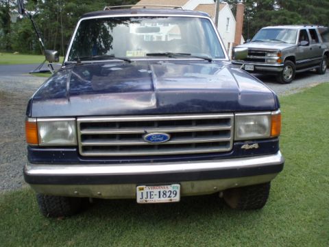 1989 Ford Bronco - Front view of a blue 1989 Ford Bronco, showing its chrome grille and bumper. Another SUV is visible in the background.