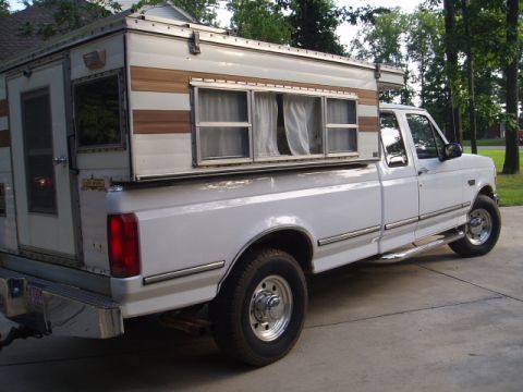 1980 Ford Bronco - The rear passenger side of a white pickup truck with a camper shell is shown parked on a concrete driveway. The camper shell has wood-grain…