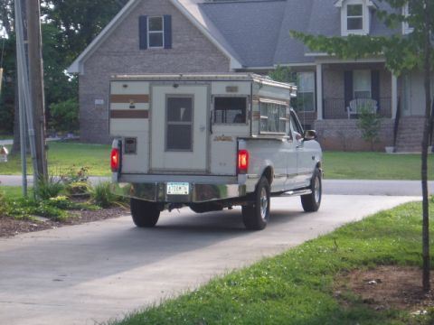 1980 Ford Bronco - The rear-left side of a white pickup truck with a camper shell is shown parked on a driveway. The truck has a chrome bumper with a license…