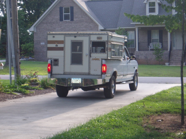 White pickup truck with camper shell, rear-left view, parked on a driveway.
