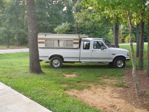 1980 Ford Bronco - A white Ford F-Series pickup truck with a camper shell is parked on a grassy area next to a tree. Another dark-colored vehicle is parked be…