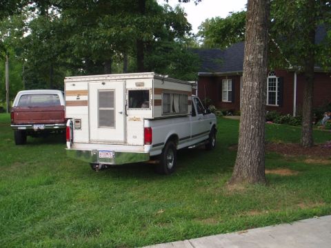 1980 Ford Bronco - A white Ford pickup truck with a camper shell is parked in a grassy yard next to a brick house. A brown Ford Bronco is parked behind it.