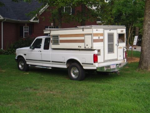 1980 Ford Bronco - A white Ford pickup truck with a camper shell is parked on a grassy lawn in front of a brick house. The truck has chrome accents and a trai…