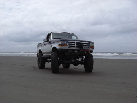 1993 Ford Bronco - This lifted pickup truck, likely a 1993 Ford Bronco, is parked on a sandy beach with the ocean in the background.
