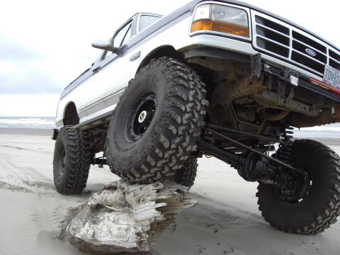 1993 Ford Bronco - Modified Ford F-Series truck navigates a beach, with its front right tire resting on a large piece of driftwood.
