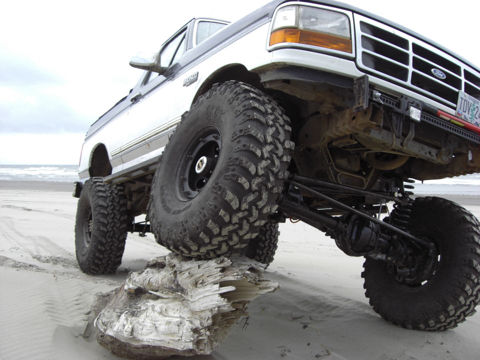 Two-tone lifted Ford F-Series truck with large off-road tires on a sandy beach, front right tire on driftwood