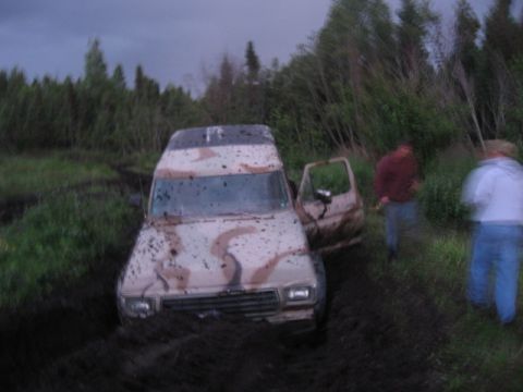 1990 Ford F-250 - A van is stuck in a muddy, wooded area with two people standing around it.