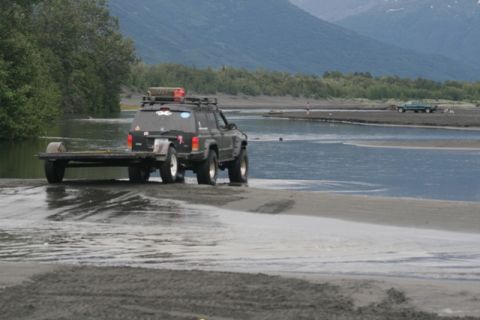 1990 Chevrolet Suburban - A black Jeep Cherokee XJ navigates shallow water while towing a trailer, with another vehicle visible in the distance.