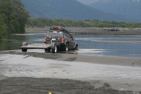 1990 Chevrolet Suburban - Jeep Cherokee towing a trailer through a river with a truck in the background.