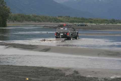 1990 Chevrolet Suburban - A Jeep Cherokee XJ navigates a shallow riverbed, with another vehicle visible in the distance.