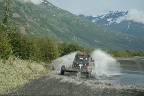 1990 Chevrolet Suburban - An SUV tows a flatbed trailer through a shallow body of water, kicking up spray against a backdrop of lush green mountains.