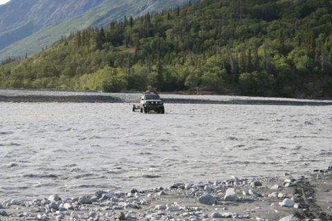 1990 Chevrolet Suburban - An SUV tows a trailer through a shallow, fast-moving river in a mountainous, forested area.