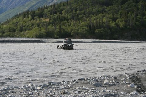 1990 Chevrolet Suburban - A black SUV navigates a shallow, rocky river with a forested mountainside behind it.