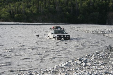 1990 Chevrolet Suburban - Off-road vehicle navigates a rocky riverbed, with a forest visible on the far bank.