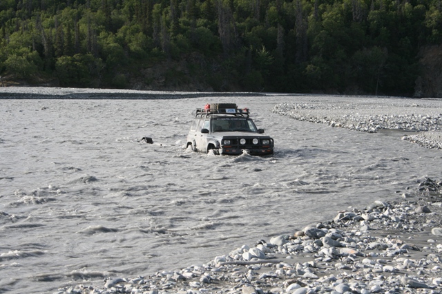 Off-road vehicle with roof rack driving through a shallow river with trees in background