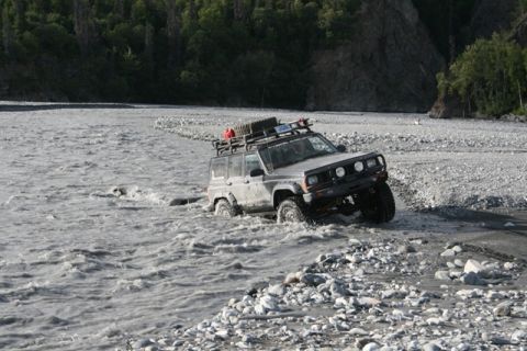 1990 Chevrolet Suburban - This modified Jeep Cherokee XJ navigates a shallow, rocky river, kicking up water.