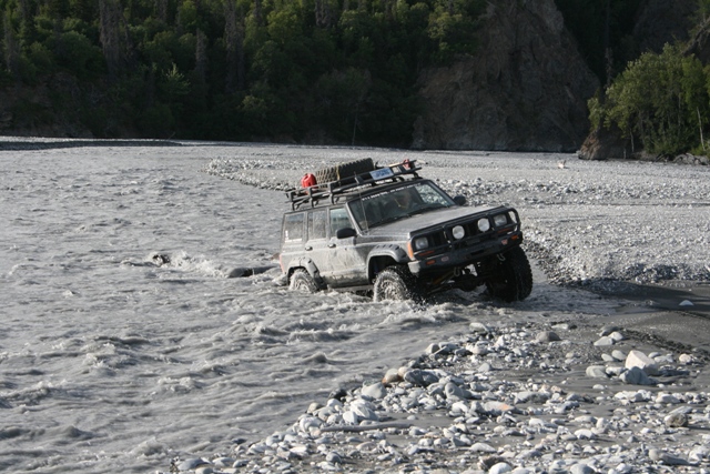 Jeep Cherokee XJ driving through a rocky riverbed with a roof rack loaded with gear