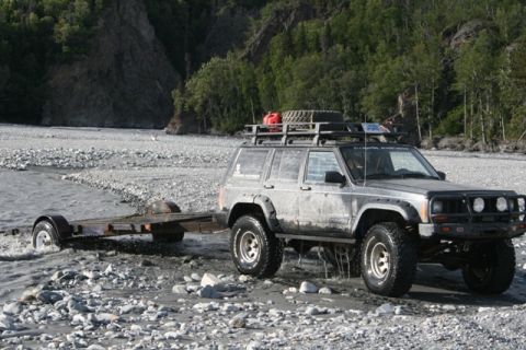 1990 Chevrolet Suburban - A muddy Jeep Cherokee XJ navigates a rocky stream bed while towing a trailer.