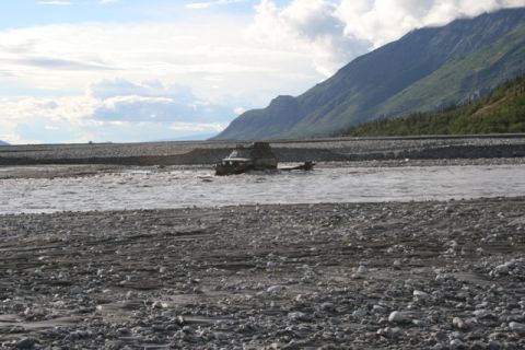 1990 Chevrolet Suburban - An off-road vehicle navigates a shallow, rocky river, with a mountainous landscape visible behind it.