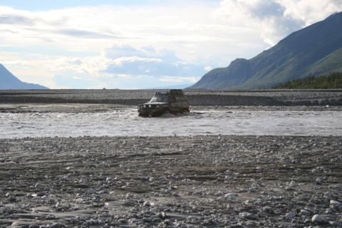 1990 Chevrolet Suburban - SUV navigates a river in a mountainous, rocky landscape under a cloudy sky.