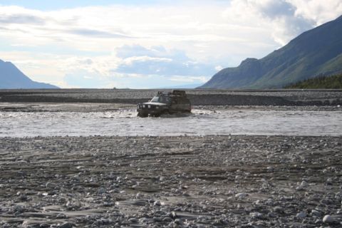 1990 Chevrolet Suburban - An off-road vehicle navigates a wide, shallow river under a cloudy sky with mountains in the distance.