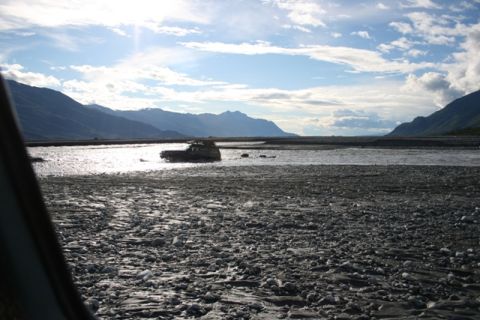1990 Chevrolet Suburban - SUV navigates a rocky riverbed under a partly cloudy sky with mountain ranges in the distance.