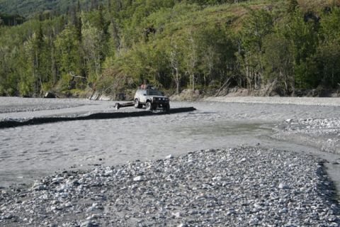 1990 Chevrolet Suburban - SUV towing a trailer through a shallow riverbed with trees in the background.