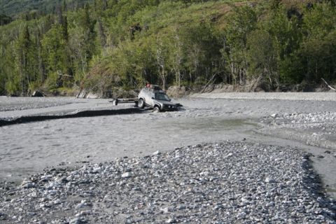 1990 Chevrolet Suburban - A four-wheel-drive vehicle navigates a rocky riverbed, pulling a small trailer.