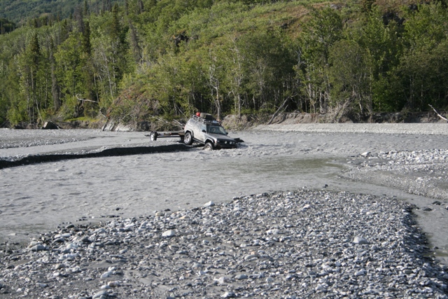 4x4 vehicle with trailer stuck in a shallow river, trees and rocky banks in background