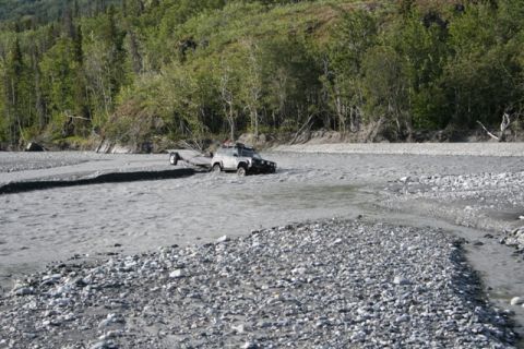 1990 Chevrolet Suburban - A gray SUV tows a trailer through a shallow, rocky river, with a dense forest lining the bank.