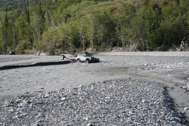 Gray SUV towing a trailer through a rocky riverbed with trees in the background