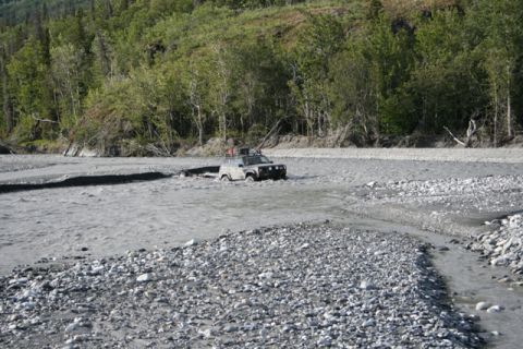 1990 Chevrolet Suburban - A gray SUV navigates a rocky riverbed, with trees lining the banks in the distance.