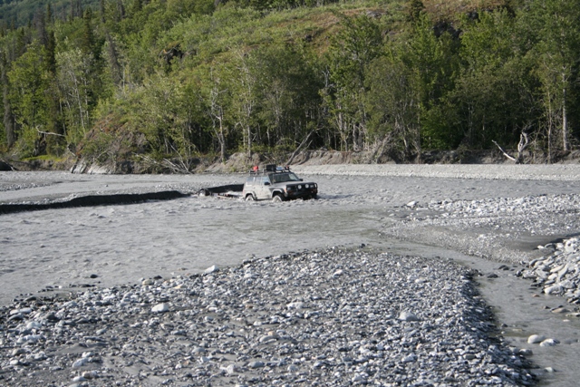 Gray SUV driving through a shallow riverbed with trees in the background