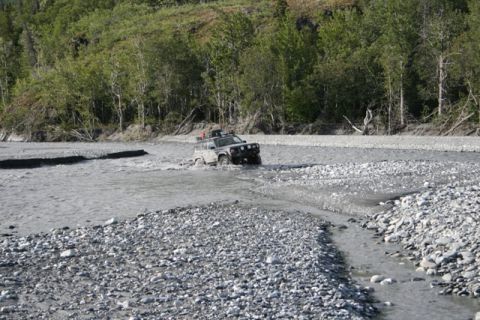 1990 Chevrolet Suburban - Off-road vehicle navigates a shallow, rocky river with trees lining the bank.