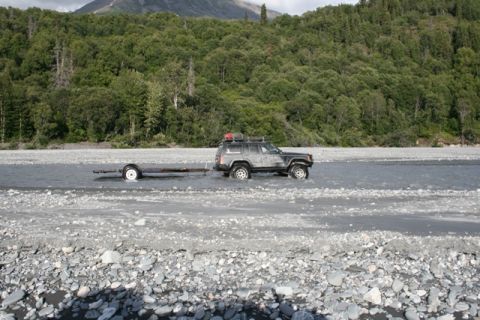 1990 Chevrolet Suburban - A Jeep Cherokee navigates a rocky riverbed while towing a trailer, with a dense forest and mountain range behind it.