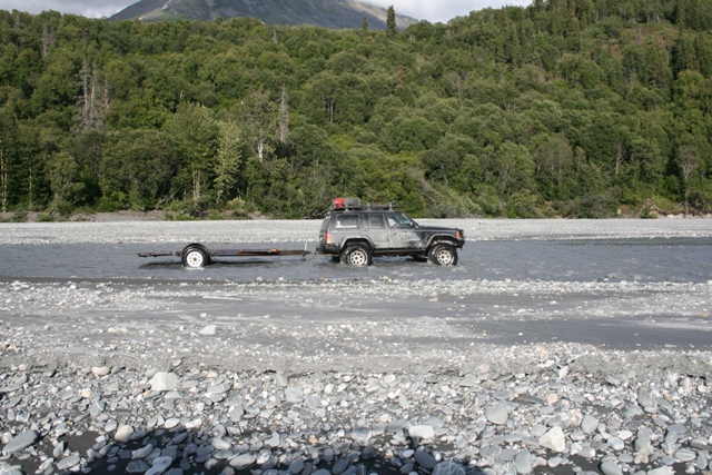 Jeep Cherokee with roof rack towing a small trailer through shallow river water, trees and mountain in background