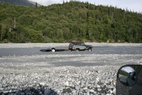 1990 Chevrolet Suburban - A dark SUV is being driven through a river while towing a trailer, with a forested mountainside behind it.