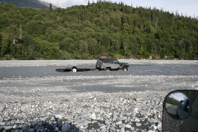 Dark SUV towing a trailer through shallow river water, with trees and mountains in the background
