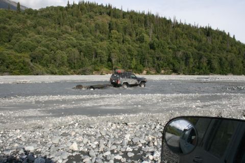 1990 Chevrolet Suburban - A dark SUV tows a trailer through a rocky riverbed, with a reflection of another vehicle visible in the foreground mirror.