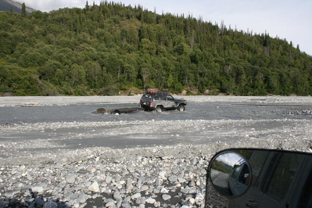 Dark SUV towing trailer through riverbed, seen from another vehicle's side mirror