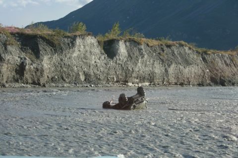 1990 Chevrolet Suburban - An off-road vehicle is submerged and partially flipped in a river, with a steep, eroded embankment behind it.