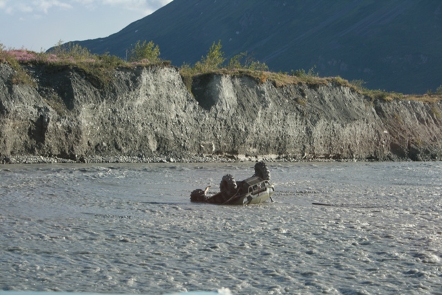 Off-road vehicle flipped over and submerged in a fast-moving river with a steep, rocky embankment in the background