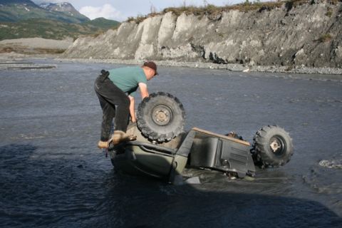1990 Chevrolet Suburban - A man struggles to right an overturned ATV in a shallow river with mountains in the background.